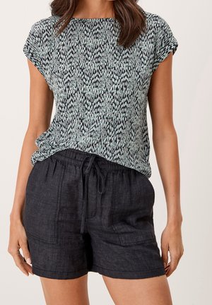 Nainen käyttää mustavalkoista kuviollista lyhythihasta puseroa, joka on sullottu mustiin nyörillisiin pellavashortseihin, neutraalia taustaa vasten.