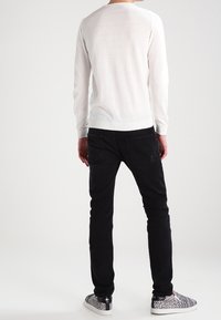 Homme portant une chemise blanche à manches longues, un jean noir et des chaussures à enfiler noires et blanches à motif, debout de dos sur un fond blanc.