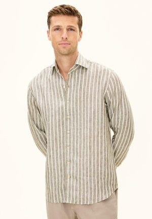 Mann mit hellbraunen Haaren, der ein beiges und weiß gestreiftes Hemd mit Knöpfen und hellbeige Hose trägt, steht mit den Händen auf dem Rücken.