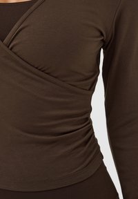 Chemise marron à manches longues avec devant style portefeuille et fronces sur le côté, portée sur le haut du torse et le bras d'une personne.