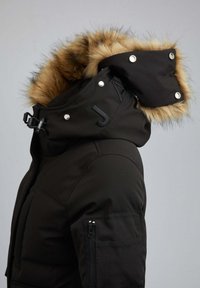 Sort parka-jakke med pelsforet hætte, trykknapper og en lynlåslomme på ærmet. Fremstillet af vandtæt stof med mat finish.