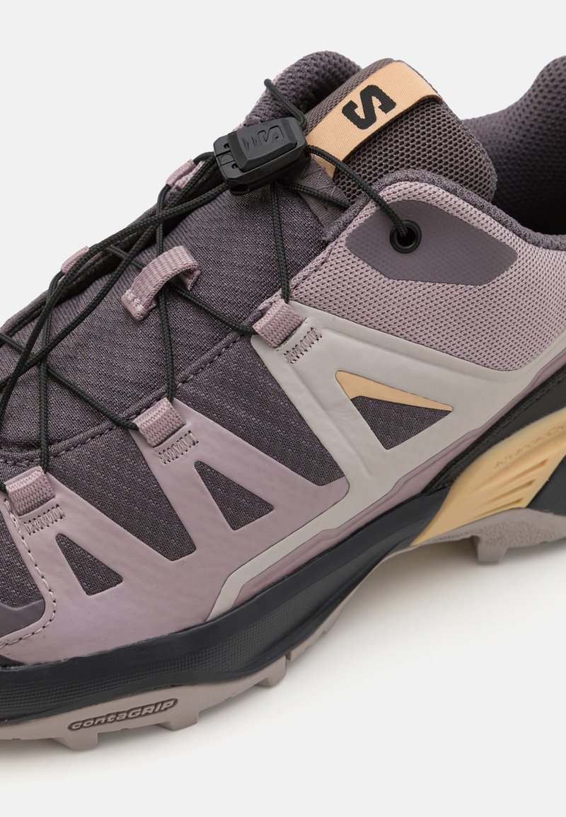 Zapatilla deportiva en tonos morado y gris, con parte superior de malla texturizada, refuerzos, sistema de cordones rápidos y suela de goma duradera.