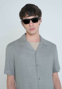 Camisa de botón de manga corta, de color gris claro y con una textura tejida, que presenta un cuello tipo camp y dos botones en el frente. Unas gafas de sol negras complementan el look.