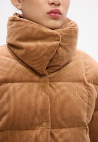 Brązowa kurtka puchowa z korduroyu z wysokim kołnierzem, z poziomymi pikowanymi panelami i wyszywanką z logo. Gładka faktura, napy.