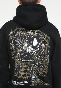 Black hoodie featuring a detailed graphic print of a character in white and gold, surrounded by spider web designs. The fabric appears textured.