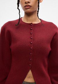 Μπορντό μάλλινο πουλόβερ με στρογγυλή λαι�μόκοψη, μακριά μανίκια και κλείσιμο με κουμπιά στο μπροστινό μέρος.