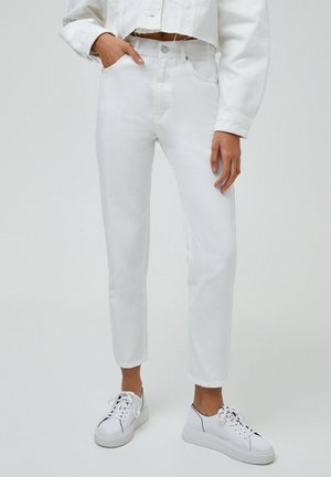 Personne portant un jean blanc taille haute raccourci, associé à des baskets blanches et une veste courte blanche, debout avec une main dans la poche.