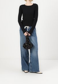 Schwarzes Langarmoberteil, lässige hellblaue weit geschnittene Jeans, hält eine geraffte schwarze Tasche mit Blumenmuster. Spitz zulaufende Schuhe.