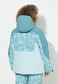 Tyrkysová nepromokavá bunda s kapucí, s bílým květinovým vzorem na zadní straně a hladkým, lehkým materiálem.