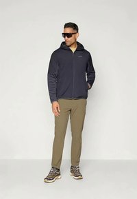 Vyras, vilkintis tamsiai pilką zip-up gobtuvą, alyvinių spalvų kelnes, žygių batus ir saulės akinius, stovi prie šviesaus vientiso fono.