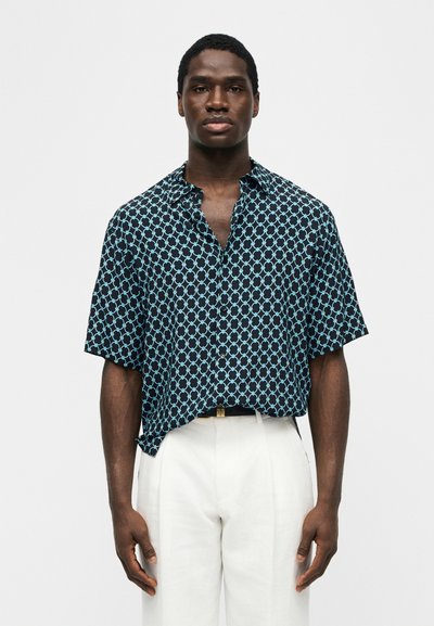 Μαύρος άνδρας που φοράει μαύρο κοντομάνικο πουκάμισο με ανοιχτό μπλε γεωμετρικό σχέδιο και λευκό παντελόνι, στέκεται μπροστά σε μια απλή φόντο.