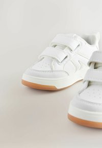 Baskets blanches de sport avec des sangles velcro, présentant une tige en cuir lisse, un bout perforé et une semelle en caoutchouc naturel.
