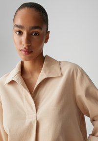 Beige collared shirt with a textured fabric. Features a buttonless placket and a relaxed fit. Smooth, soft texture without patterns.