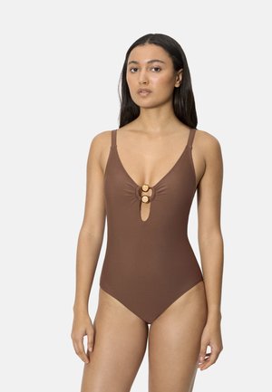Mujer con traje de baño de una pieza marrón con tirantes delgados y dos botones decorativos redondos en el detalle de abertura frontal, posando contra un fondo liso.