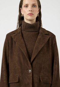 Ruskea mokkainen bleiseri, jossa on lovikaulus, yksittäinen nappisuljenta ja sivutaskut, kerrospukeutuneena ruskean ribbineuleisen pooloneuleen päälle.