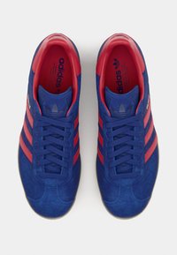 Et par blå Adidas sneakers med røde striber og rød inderside, set ovenfra, snøret op og med sidetekst "Gazole".