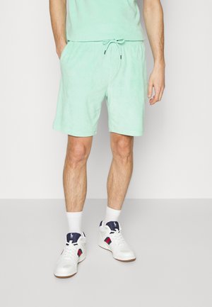 Mann trägt hellgrüne Shorts mit Kordelzug, weiße Socken und weiße Sneakers mit marineblauen und roten Details, steht vor weißem Hintergrund.