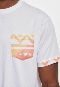 White short-sleeve t-shirt featuring a patterned pocket in orange and yellow hues, with geometric designs and accents on the sleeve.