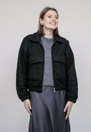 Veste zippée noire texturée avec un col, dotée de deux poches avant. Portée sur un pull gris et une jupe grise lisse.