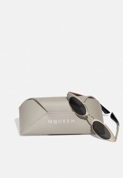 Owalne okulary przeciwsłoneczne z ciemnymi soczewkami i złotą metalową oprawą obok jasnobeżowego, teksturowanego etui z wytłoczonym logo „MQUEEN”.
