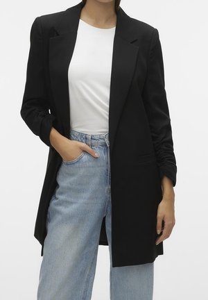 Vrouw met een zwarte blazer over een wit shirt en lichtblauwe jeans, met één hand in haar zak, staand tegen een effen achtergrond.