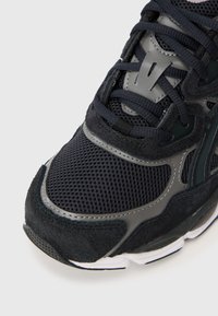 Svart atletisk sneaker som har mesh- och mockamaterial, en dämpad sula och grå accenter. Inkluderar robusta snören och en texturerad yta.