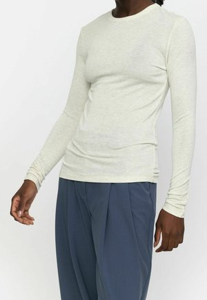 Personne portant une chemise ajustée à manches longues de couleur beige clair et un pantalon large bleu foncé plissé, debout devant un fond blanc.