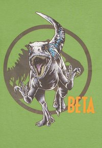 Camiseta de algodón verde con un gráfico de un dinosaurio rugiente, etiquetado como "BETA" en naranja, con un fondo de silueta circular oscura.