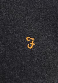 Tissu gris foncé avec une petite lettre minuscule « f » stylisée brodée en fil orange près du centre.