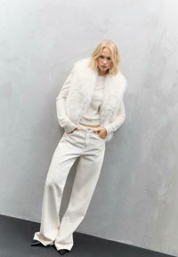 Weißer Kunstpelz-Weste über einem hellen Pullover, kombiniert mit cremefarbenen, weit geschnittenen Hosen und schwarzen, spitz zulaufenden Schuhen vor einem neutralen Hintergrund.