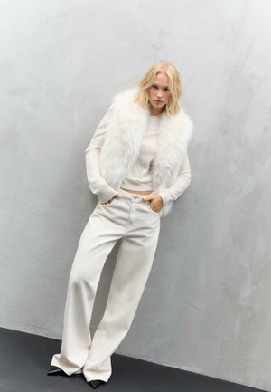 Gilet in pelliccia sintetica bianca sopra un maglione chiaro, abbinato a pantaloni a gamba larga crema e scarpe nere a punta su uno sfondo neutro.