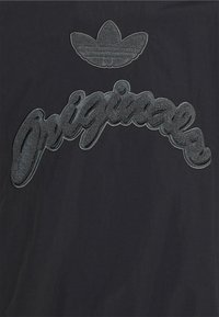 Svart t-shirt med en texturerad, broderad "Originals"-logotyp i mörkgrått och en Adidas trefoil-emblem ovanför.