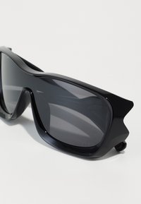 Black sunglasses with a sporty design, featuring large, dark lenses and angular frames. Accented with textured detailing on the temples.