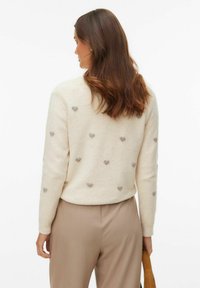 Pull de couleur crème orné de motifs en forme de cœurs gris dispersés, fabriqué dans un matériau doux et texturé, associé à un pantalon beige.
