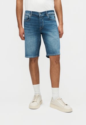 Άνδρας που φοράει μπλε τζιν σορτς, λευκές κάλτσες μέχρι τον αστράγαλο και εκρού αθλητικά παπούτσια, στέκεται μπροστά σε ένα απλό φωτεινό φόντο.