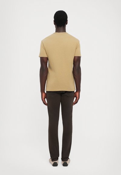 Μακριμάνικη, μπεζ βαμβακερή μπλούζα με στρογγυλή λαιμόκοψη, συνδυασμένη με καφέ, εφαρμοστά παντελόνια και ανοιχτόχρωμα παπούτσια. Απλό φόντο.