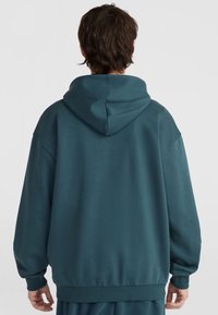 Turkizna kapuca s širokim krojem, rebrastim manžetama i rubom, s gladkom, teksturiranom tkaninom i jednostavnim dizajnom.