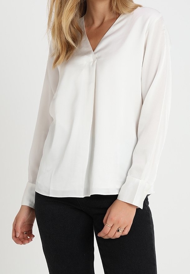Femme portant un chemisier blanc à manches longues avec un col en V et un détail plissé à l'avant, ainsi qu'un pantalon noir, visible des épaules aux hanches.