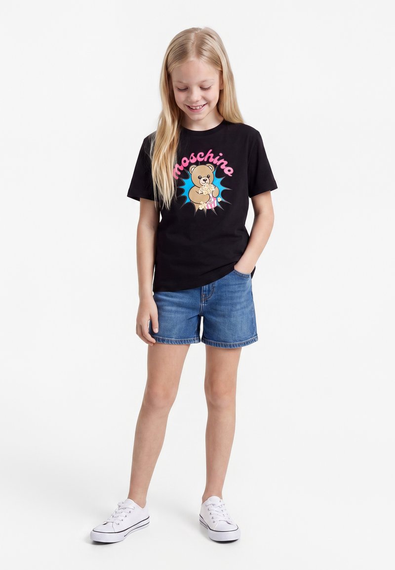 Млада момиче, облечена в черна тениска Moschino с графика на плюшено мече, сини дънкови къси панталони и бели маратонки, стои с една ръка в джоба.