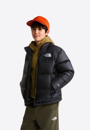 Jaunas asmuo dėvi oranžinę kepurę, juodą pūkinę striukę, alyvuogių žalią džemperį su gaubtu ir atitinkančias kelnes su The North Face logotipais.