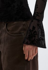 Črna prosojna bluza z vzorcem iz žameta v cvetličnem dizajnu, razširjene rokave, kombinirana s rjavimi hlačami iz korduroja z vidnim žepom in kovinskim gumbom.