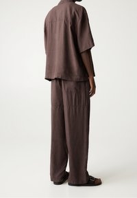 Osoba ubrana w luźną, brązową koszulę z krótkim rękawem oraz dopasowane szerokie spodnie, w zestawieniu z czarnymi sandałami na tle jednolitego jasnego tła.