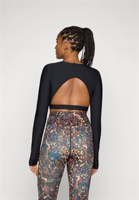 Svart langermet topp med et stort ovalt utsnitt på baksiden, sammen med flerfargede, mønstrede høytaljede leggings med mesh-detaljer.
