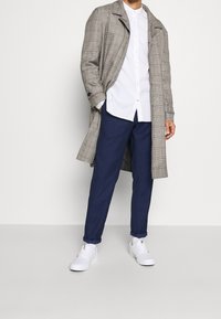 Trench coat a quadri grigi, camicia bianca con bottoni, pantaloni blu scuro e sneakers bianche. Il cappotto presenta un colletto classico e una lunghezza lunga.