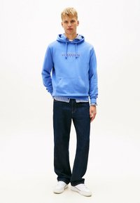 Felpa blu chiaro con tasca frontale e logo, indossata sopra una maglietta a righe, abbinata a jeans scuri e sneakers bianche.