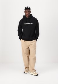 Hanorac negru cu logo alb "AIR", pantaloni bej, lejeri, și adidași ușori. Modelul stă în fața unui fundal alb.