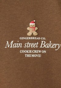 Tessuto marrone con testo bianco: "GINGERBREAD CO. Panetteria di Main Street COOKIE CREW IN MOVIMENTO!" con un grafico di un omino di pan di zenzero che indossa un cappello di Babbo Natale.