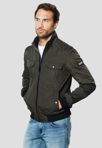 Kaki krāsas bumbvedēja jaka ar diviem krūšu kabatām, rāvējslēdzēja aizdari, ribota apmali piedurknēm un apkaklei. Valkāta uz baltas krekla ar ziliem džinsiem. Gluda tekstūra.