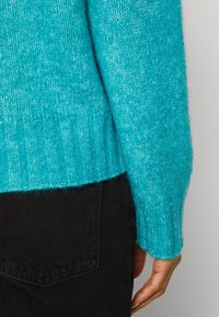 Turkos stickad tröja med ribbad nederkant och manschetter, tillverkad av mjukt, texturerat material. Kombineras med svarta jeansbyxor.