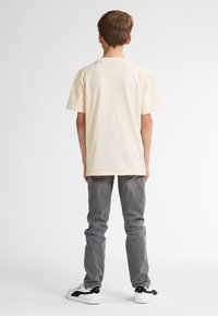 Beige katoenen T-shirt met korte mouwen, gecombineerd met grijze spijkerbroek en witte sneakers met zwarte accenten, gezien van achteren.